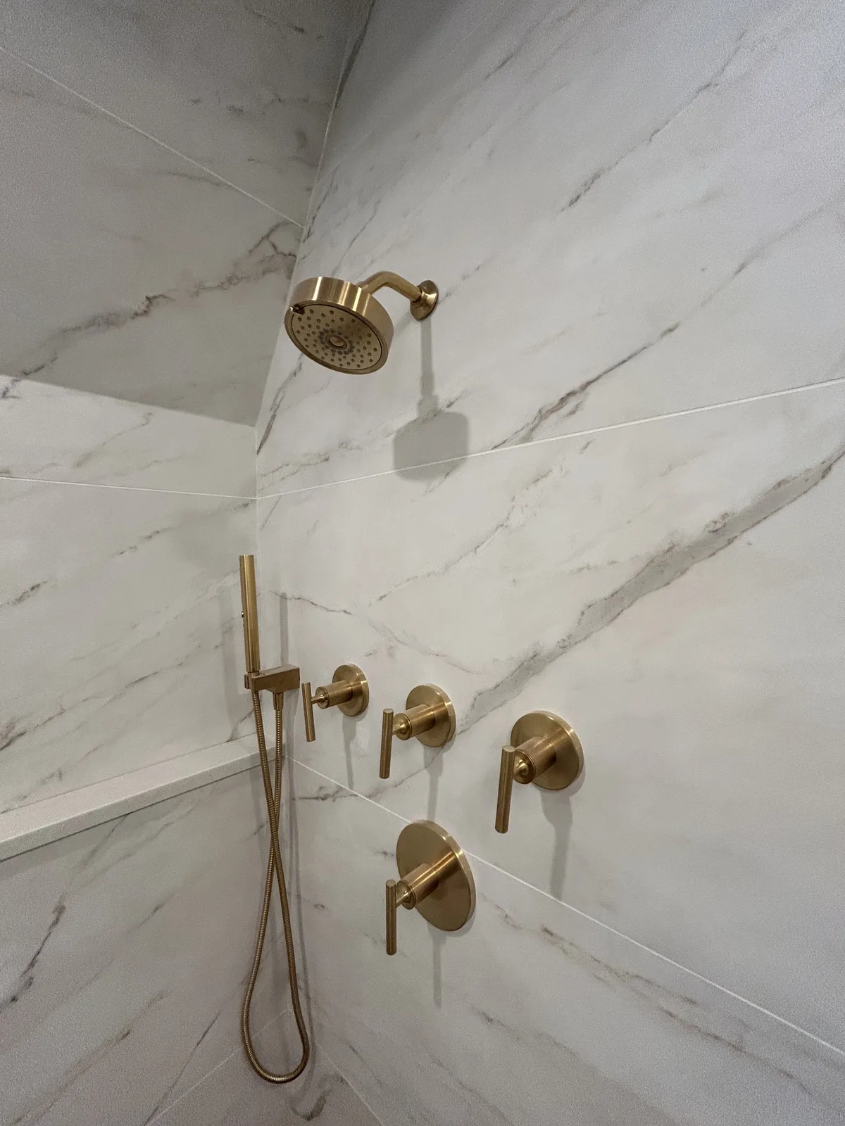 Bookmatched marble-look shower wall with brushed-brass rain head, hand shower, and trim